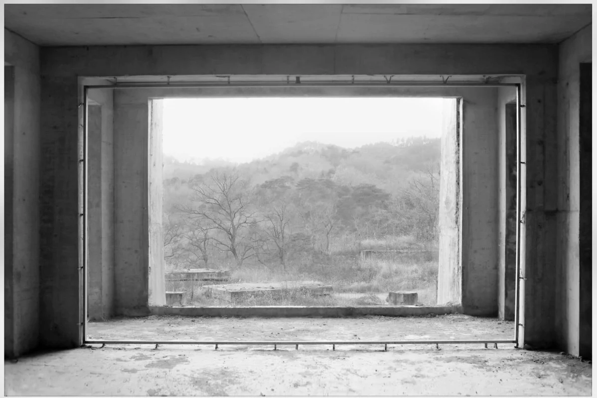 View through a large window opening in an unfinished building framing trees and mountains outside