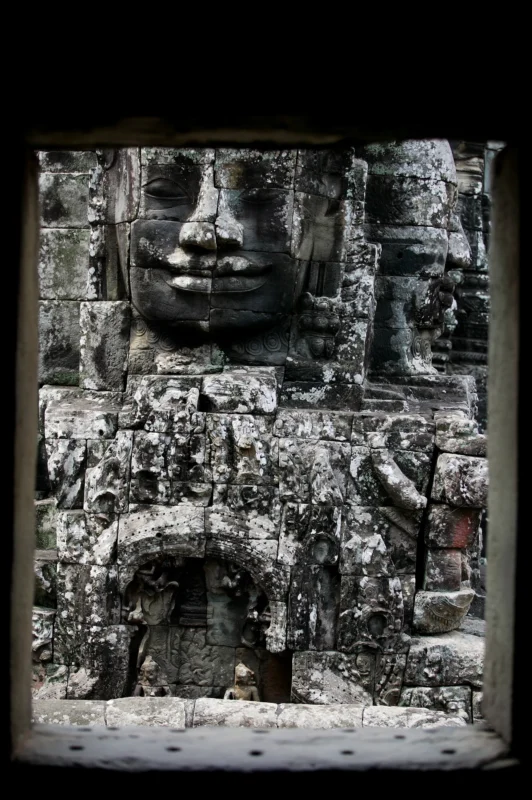 A monumental stone face emerging from the weathered ruins of Angkor, surrounded by intricate carvings and centuries of erosion.