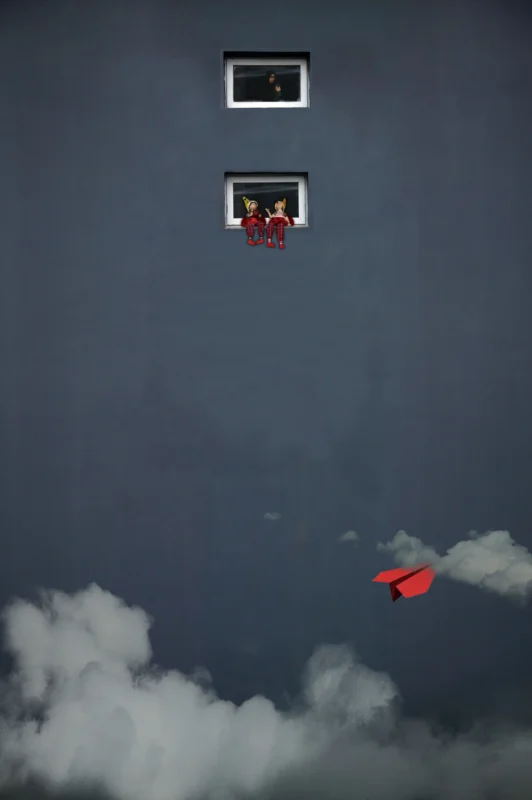 A red object floating beneath a small window against a grey wall.