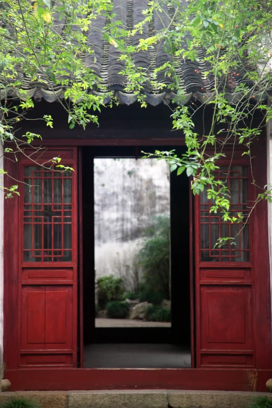A red doorway opening toward a misty garden framed by hanging branches.