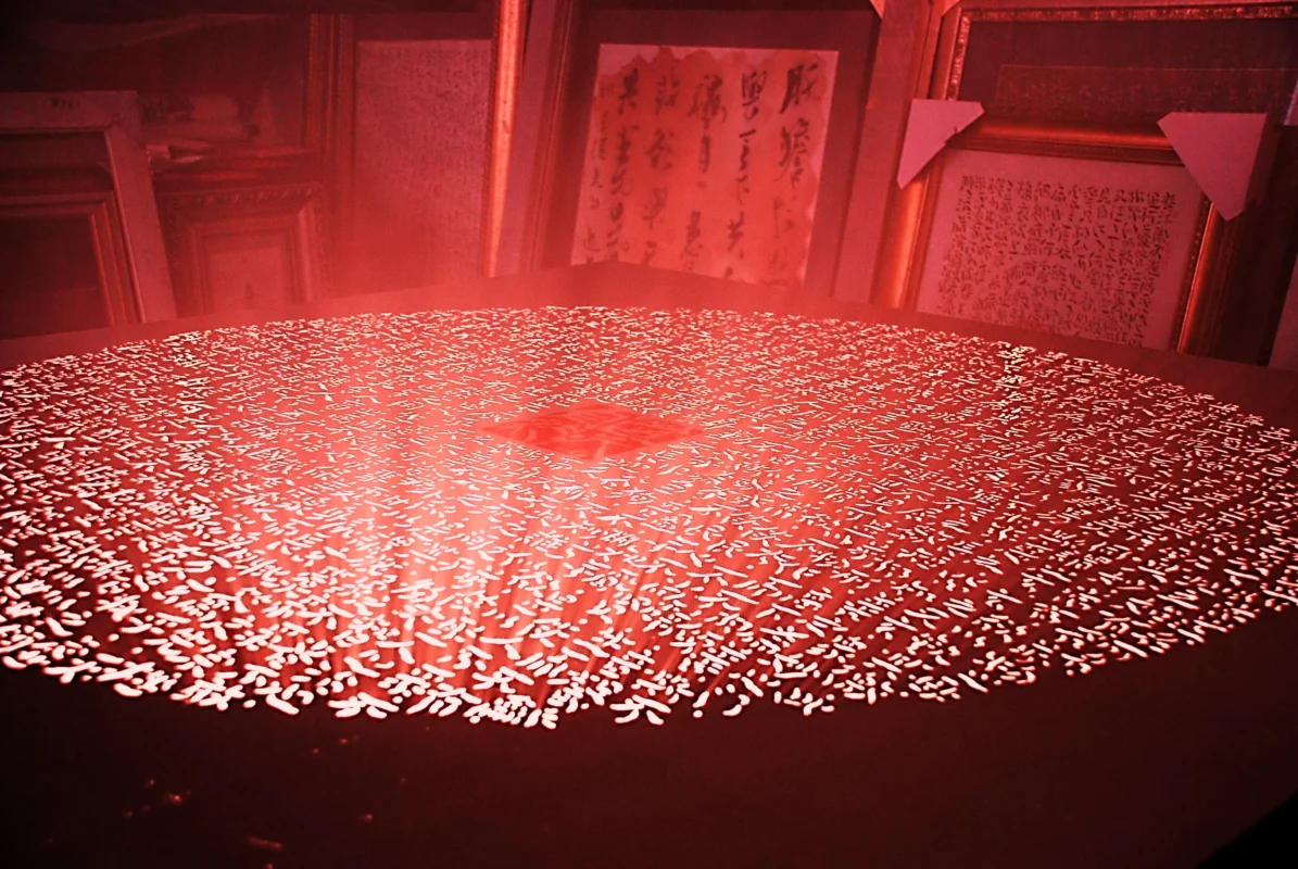 A circular field of perforated characters radiates red light, while a square seal-like image emerges at its center, creating an immersive environment of light and atmosphere.