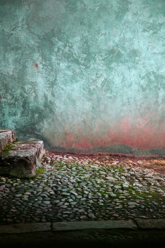 An aged turquoise wall, weathered stone steps, and a cobblestone ground create a quiet courtyard scene filled with traces of time.