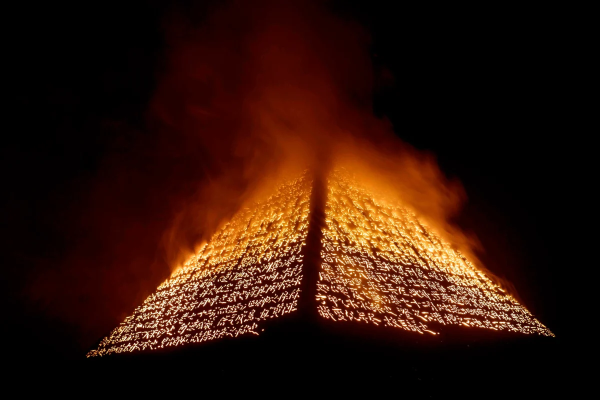A pyramid-shaped installation composed of perforated incense-trace text glows from within as dense smoke rises from its summit, creating a powerful ritualistic atmosphere.