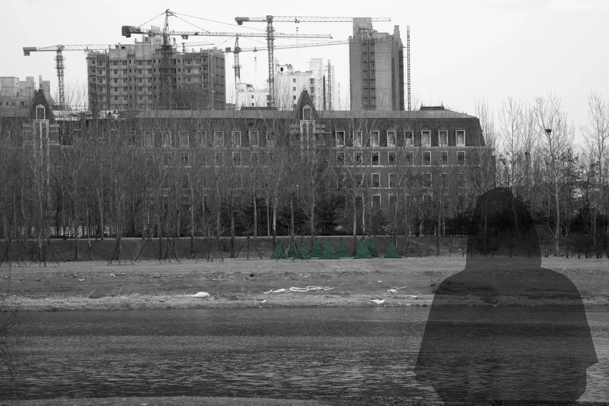 A dark transparent figure stands before an urban riverside landscape, where construction cranes and unfinished buildings rise in the distance.